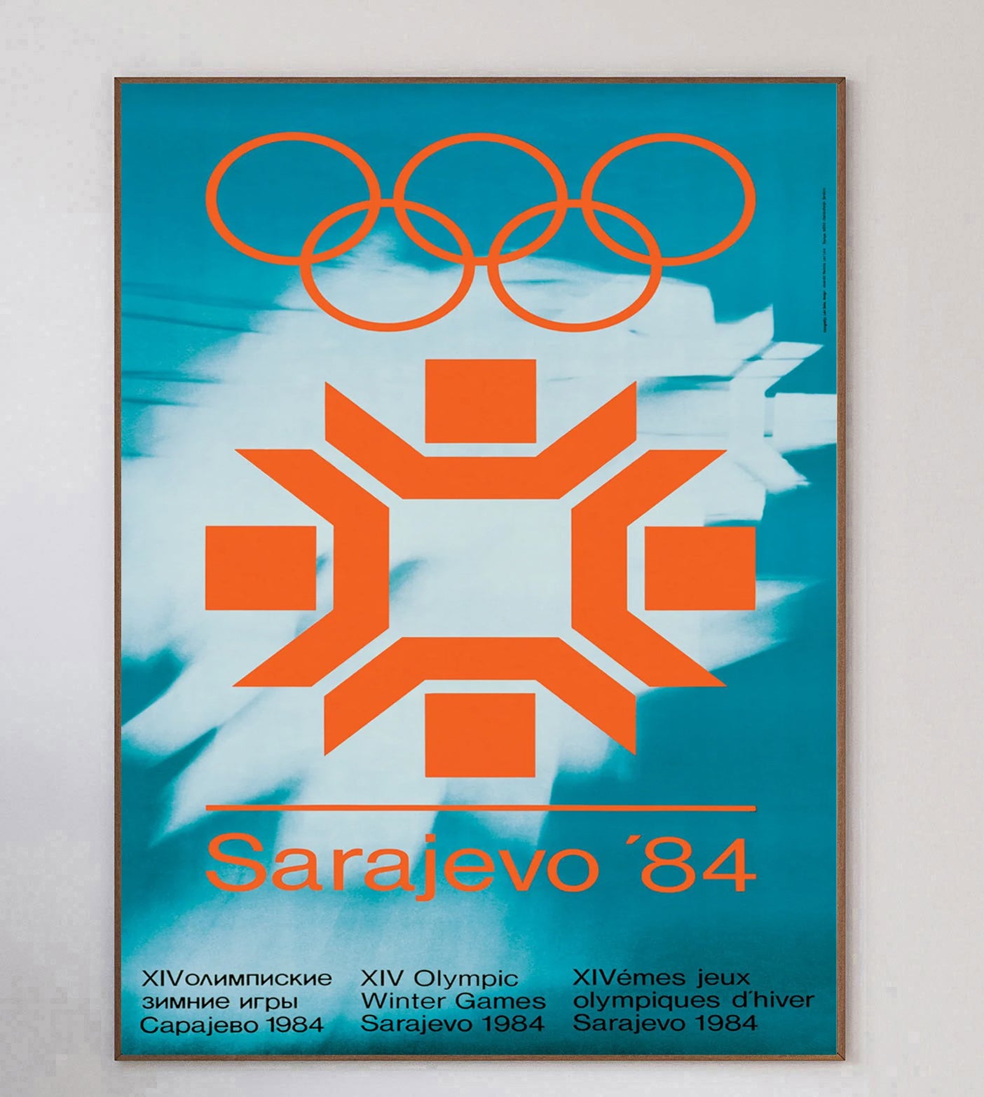 Плакат Сараево 1984: оранжевая снежинка и олимпийские кольца на бирюзовом фоне.