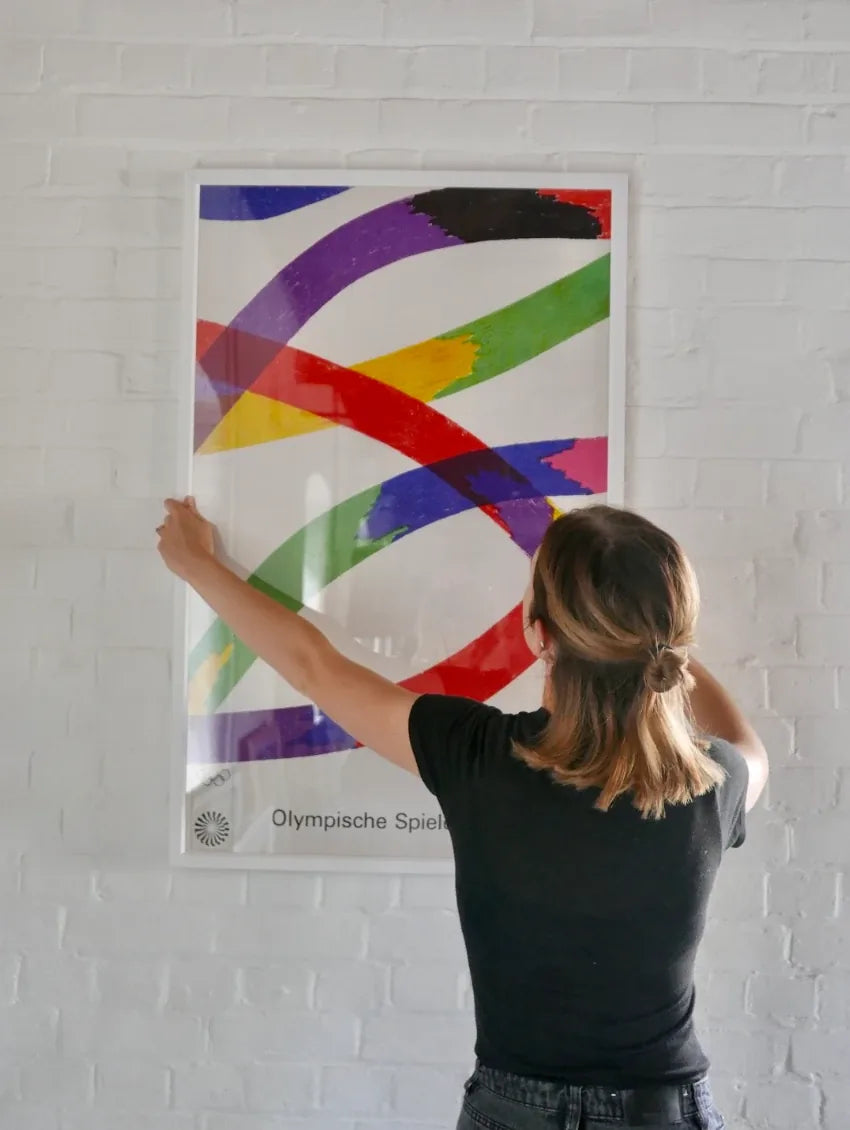 An original Munich 1972 Olympic Games poster framed to the edge in a white moulding, being hung on a brick wall